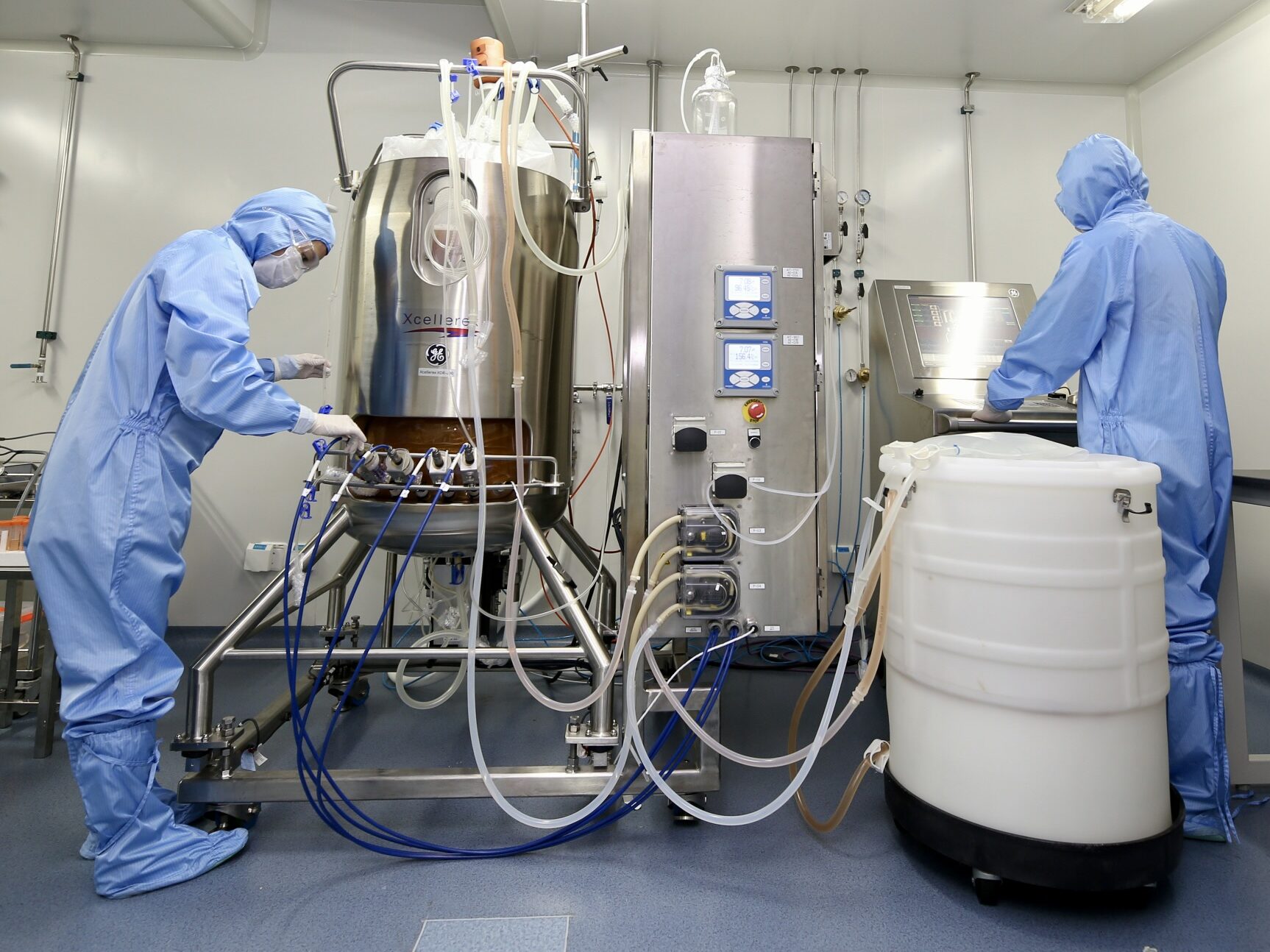 Two scientists wear blue safety suits in front of a large reactor with tubes and cords going in and out.